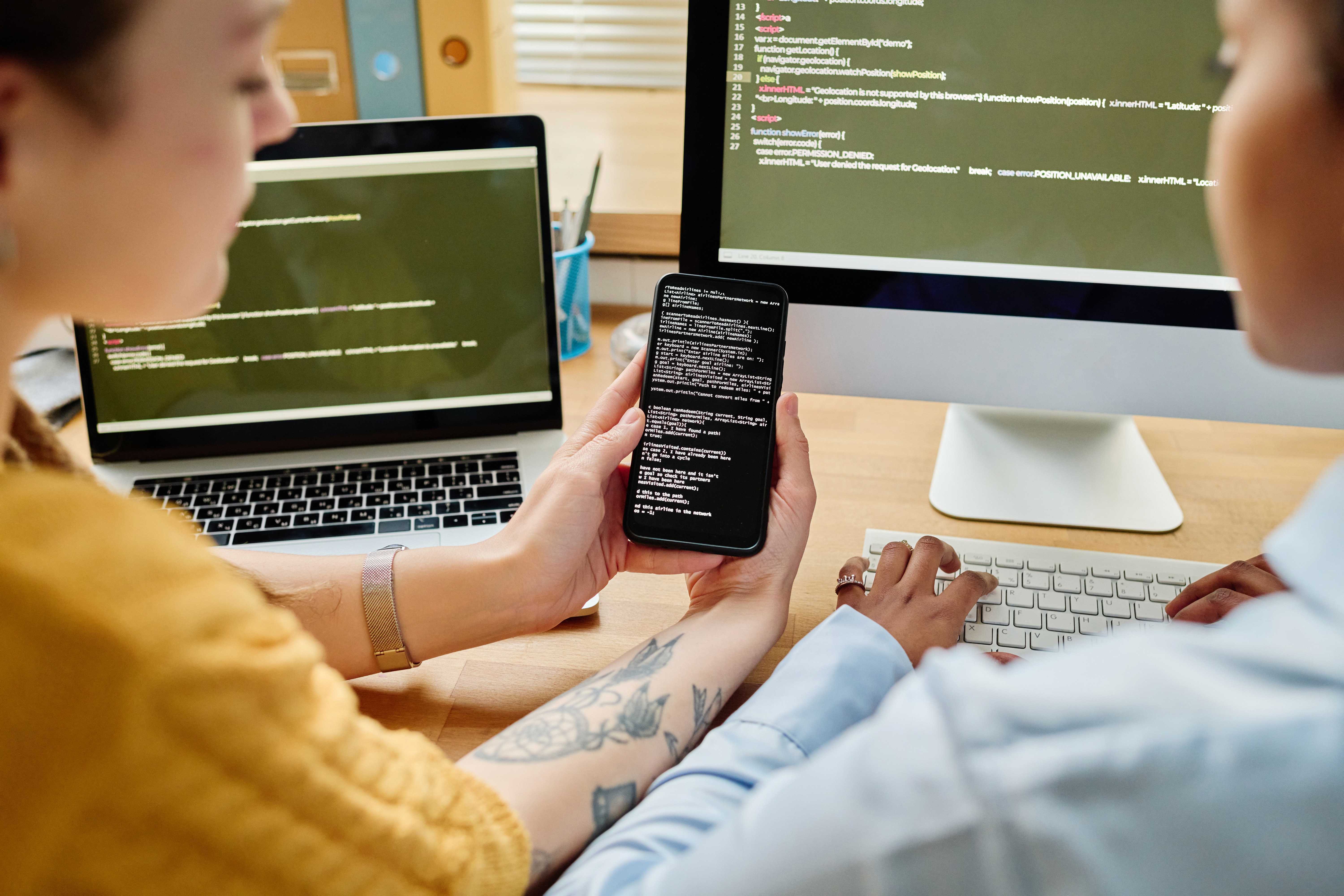 Two individuals are collaborating in a modern workspace, focusing intently on dual computer screens displaying complex code. One person types on a keyboard while the other holds a smartphone, seemingly reviewing data. The atmosphere is professional and concentrated, highlighting teamwork in a technological field. This image is ideal for illustrating topics such as software development, cybersecurity, IT solutions, or modern business operations. It could be used to convey ideas of innovation, digital transformation, and the future of work.