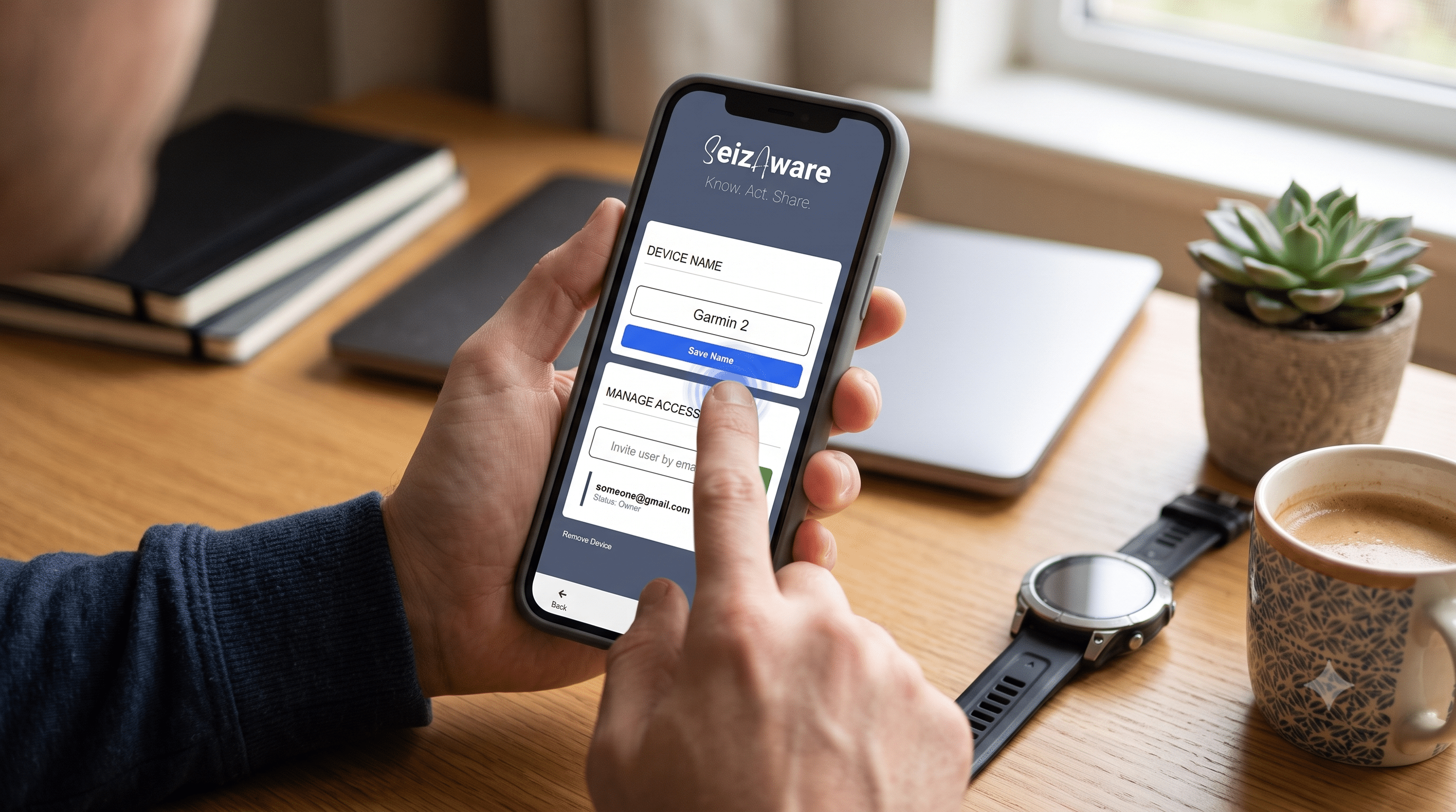 A person holding a smartphone and tapping the blue "Save Name" button in the SeizAware app, with a blurred Garmin smartwatch and coffee cup on a wooden home-office desk in the background.